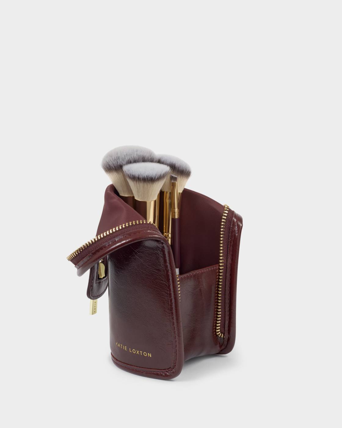 A burgundy leather makeup brush case with gold zippers and a small gold-tone brand plaque, shown open with several makeup brushes inside on a light gray background.